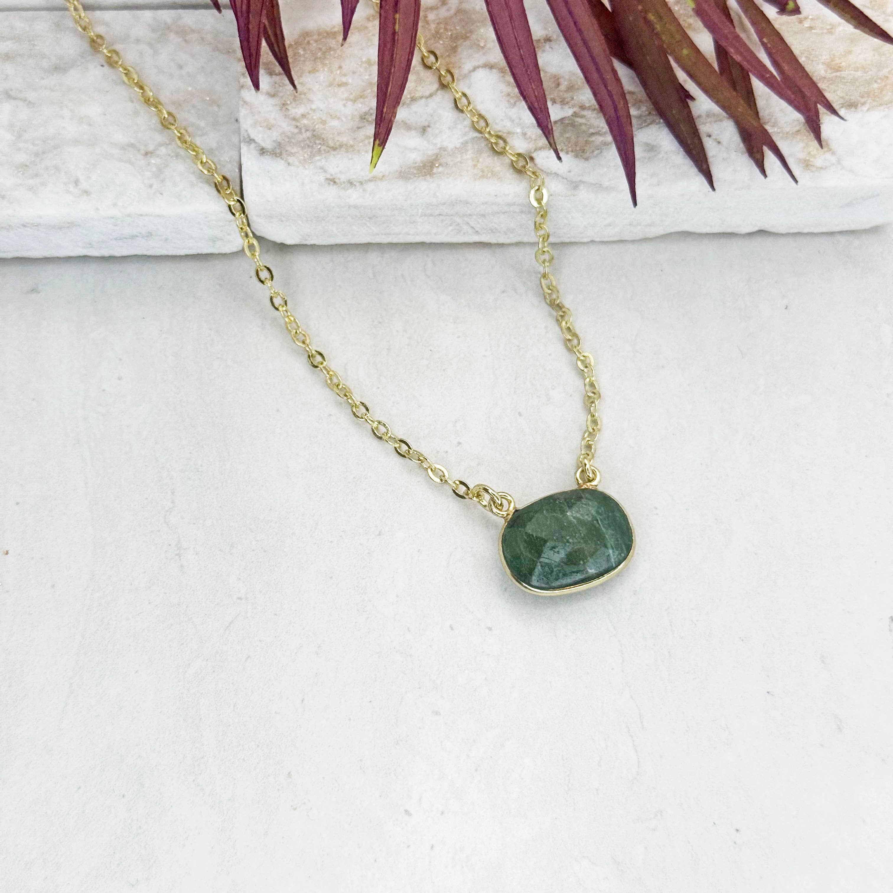 Gold necklace with a green stone pendant on a white surface with a plant in the background